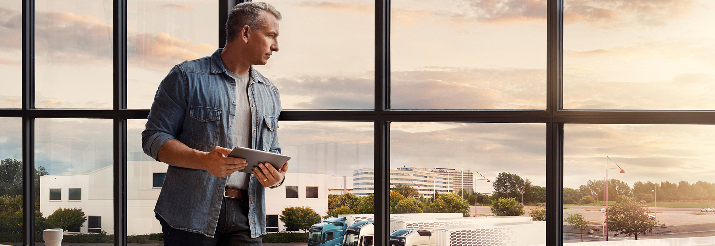 Tahvelarvutit hoidev mees seisab akna juures ja vaatab alla oma veokipargi poole